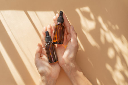Womens hands, holding two bottles of amber glass with hyaluronic serum. Unmarked packaging for cosmetics on beige background with striped shadow. Top view, flat lay, copy space.Concept of mockupの写真素材