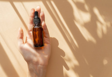 Amber glass bottle with pipette in female hands. Cosmetic serum for face, essential oil close-up on brown background. Copy space, flat lay.の写真素材