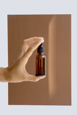 Womens hands hold bottle of amber glass with cosmetic serum, retinol on brown background. Mockup of container with dropper lid with collagen in rays of sunlightの写真素材