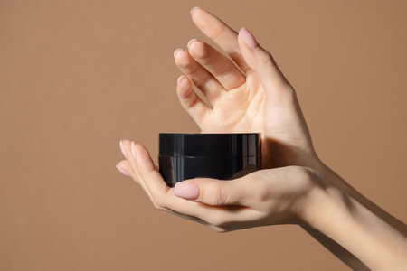 On palm of womans hand is Black jar of moisturizing cream on brown background. Mockup of bottle without labeling with an anti-aging mask, cosmetic scrub. Isolated beige background with copy spaceの写真素材