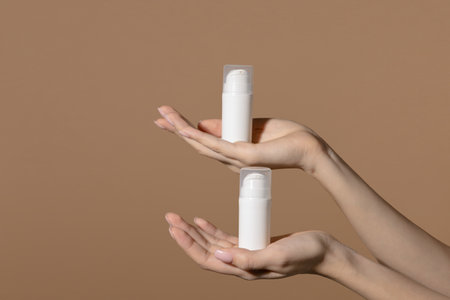 White unmarked bottles for cosmetics stand on womens well-groomed palms. Mock up of container for serum, lotion, cream, retinol. Light brown background with copy spaceの写真素材