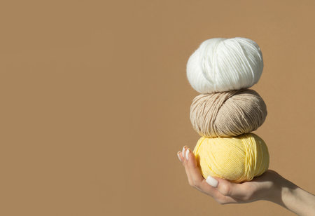 Female hand hold a stack of a large three tangles woolen thread on a light brown background. Woman holding knitting woolen threads white, yellow and brown colors. Mockup for designers, copy spaceの写真素材
