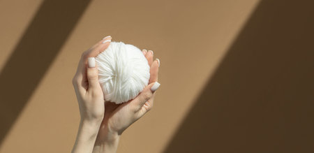 There is a white ball of knitting thread in palm of woman's hand. Hand holds a ball of yarn on beige...