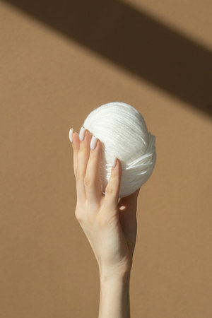 Skein of white woolen threads in women's hands on beige background. Sun's rays fall on ball. Concept of needlework, hobby.の写真素材