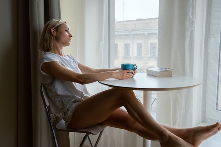 Young woman with cup of coffee is reading a book sitting home relaxing by the windowの写真素材