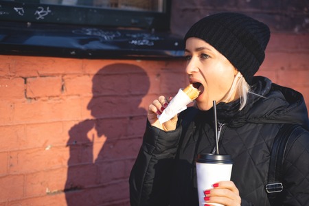 Cheerful woman in the street drinking morning coffee an d eat hot dog at winter timeの写真素材