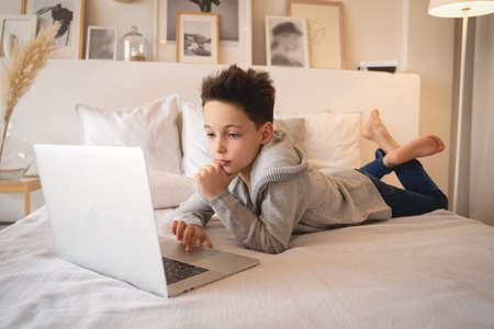 A little boy sitting on a big bed with a laptop book and looks into the camera.の写真素材