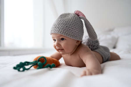 Baby four months old in a bunny costume with a knitted carrot laying on white bedの写真素材