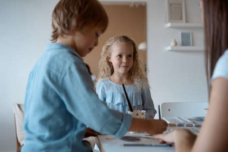 Group of kids and their teacher leaning over table with watercolor paints and painting self-made clay items. Children with teacher in art classの写真素材