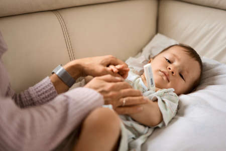Worried young mother sitting on sofa beside her sick son with high fever. Mom measures temperature using thermometer of sick child lying under blanket at homeの写真素材