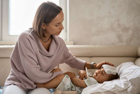 Young little boy on the sofa with cooler gel on her head and measuring temperature by thermometer. and take care by her momの写真素材