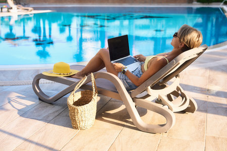 Hipster woman using laptop by the pool. Young girl freelancer working on a laptop while on vacation...