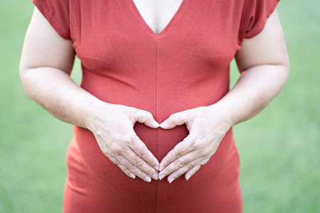 Close-up image of pregnant woman in red maternity clothes making heart shape on her baby bump.