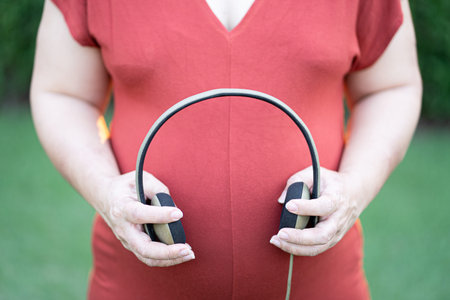 Close-up image of pregnant woman in red maternity clothes holding headphone on her baby bump.