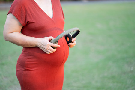 Image of pregnant woman in red maternity clothes standing in garden holding headphone on her baby...
