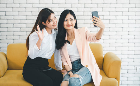 Pretty two asian woman smiling sitting at living room and taking selfie on mobile phone.の写真素材