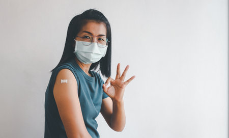 Asian woman wearing protective mask against covid-19 with giving ok hand sign to rolling out after getting vaccine.の写真素材