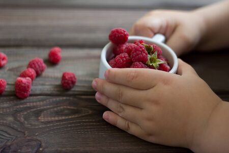 child's hands hold a cup of raspberries. Close-up. Wooden background.の写真素材