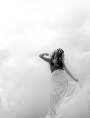Beautiful young girl in a long white skirt goes through fog, smoke and clouds outdoors.の写真素材