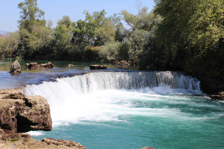a low foamy waterfall over a bright turquoise lake surrounded by rocks and trees under a clear summer skyの写真素材