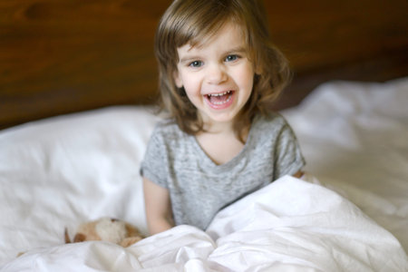 Active adorable baby wakes up lying in bed with her Teddy bear toy, soft white linens. Children's...