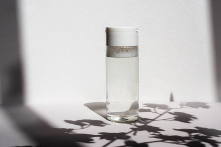 Bottle with clear micellar water on a white background with a shadow from flowers. Lotion for cleansing the skin and eyes from cosmetics, makeup. Beauty product in white plastic packaging.の写真素材