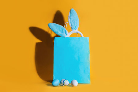 Blue shopping bag, rabbit ears and Easter eggs with a shadow on a yellow background. Online shopping...
