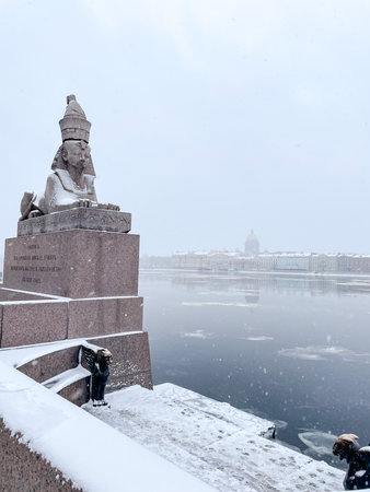 A beautifully serene winter landscape showcases a statue elegantly standing by a frozen river amid gentle snowfallの写真素材