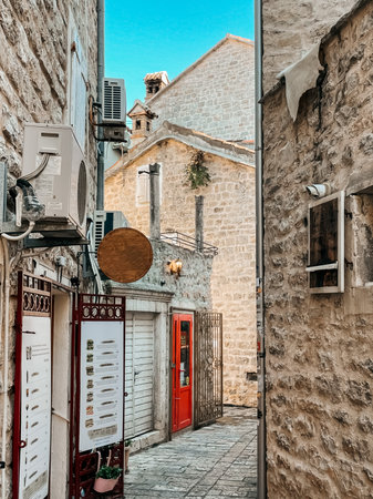 narrow stone alley with red doorway and menu signs, warm sunlight, worn cobblestone, airconditioners and textured facades creating cozy travel vibe , Budvaの写真素材