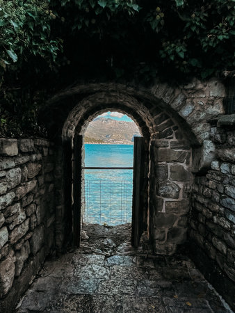 stone arch framing turquoise sea beyond, weathered masonry opens onto secluded cove, mossy stones and pebble path leading to calm horizon, moody sky casting cool tonesの写真素材