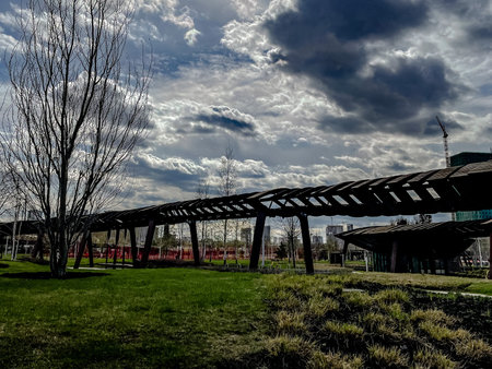dark canopy silhouette against stormy sky, lone jogger pausing under timber structure, heavy cloudscape creating moody contrast, grassy foreground, playground structures in distanceの写真素材