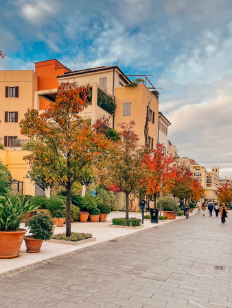 Autumn promenade lined with potted trees and colorful foliage under golden sky, shoppers and commuters strolling along wideの写真素材