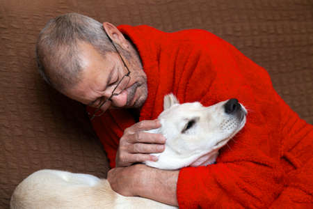 Male owner in red dressing gown lovingly hugs his white dog. High quality photoの写真素材