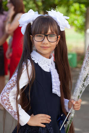 Half-length portrait of cute adorable serious thoughtful first grader girl in school uniform and white bows in long brown hair on blurred light green sunny grass background. High quality photo.の写真素材