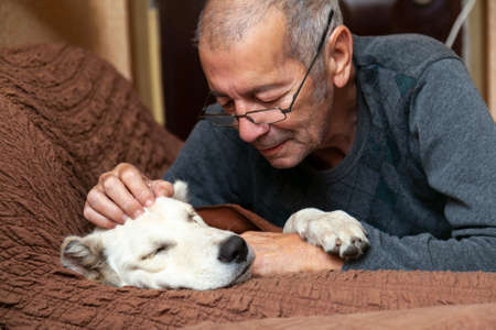 Male owner cuddling his white dog sleeping on the couch. Close-up portrait.の写真素材