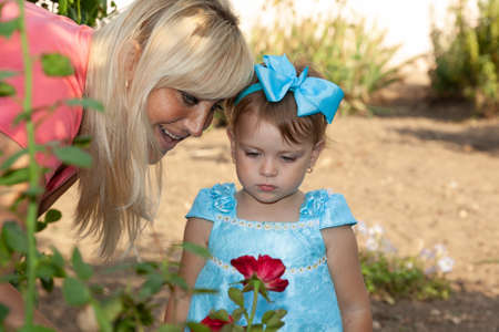 Young mother and her little daughter look at a red rose flower in the garden. High quality photoの写真素材