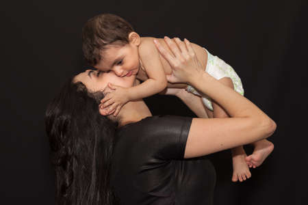 A young mother holds her little son over her head and kisses him. Isolated portrait on black background.の写真素材
