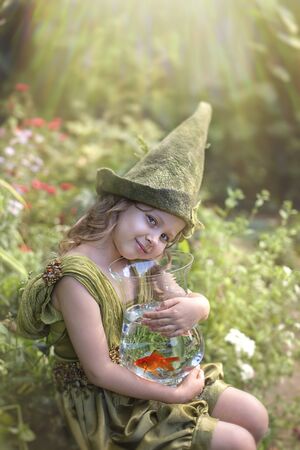 Girl in a green gnome hat hugs an aquarium with a goldfish in a green garden.の写真素材