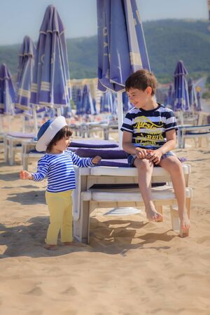 Two children in striped T-shirts are standing on the beach near sun loungersの写真素材