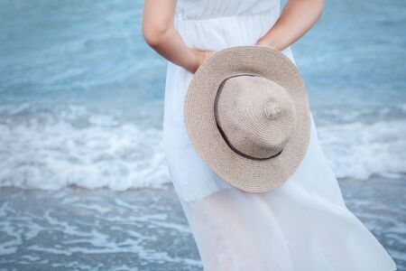 A young girl in a white dress stands with her back on the shore of the blue sea, holding a light hat behind her. Close up. Horizontalの写真素材