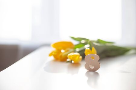 Wooden number eight stands on a white table. Behind in soft focus lies a bouquet of yellow tulips. International Women's Day . Copy spaceの写真素材