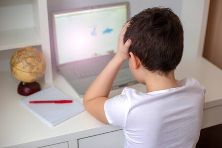 A preteen schoolboy in a white T-shirt, tired of sitting at the monitor with a laptopの写真素材