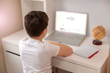 The boy sits with his back behind a laptop, looks at the monitor. Distance learning. Home Study Exam Preparationの写真素材