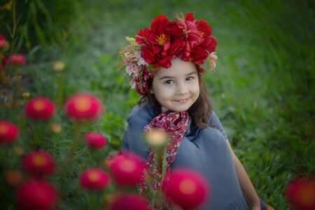A little girl with a red floral crown sits under a flowering bush with burgundy carnationsの写真素材