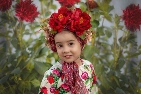 A little girl with a red floral crown sits under a flowering bush with red big flowersの写真素材