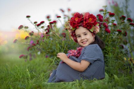 A little girl with a red floral crown sits under a flowering bush with burgundy carnationsの写真素材
