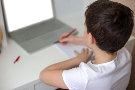 The boy sits with his back behind a laptop, writes something in a notebook
