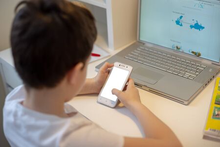 The boy sits with his back behind a laptop, holds a white smart phone in his hands, looks into it. Distance learning. Smartphone prevents learningの写真素材