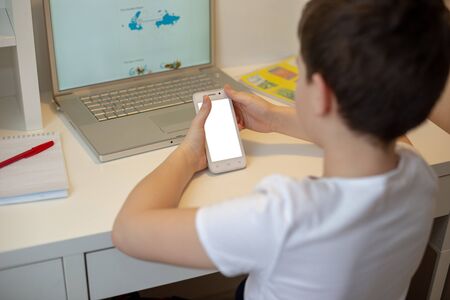 The boy sits with his back behind a laptop, holds a white smart phone in his hands, looks into it. Distance learning. Smartphone prevents learningの写真素材