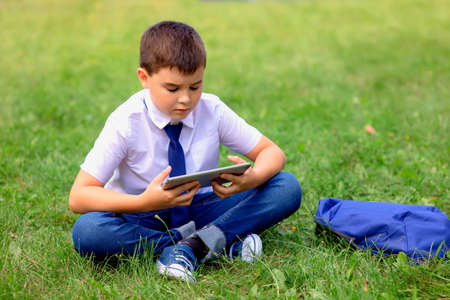 Serious Schoolboy in a white shirt and blue tie sits on green grass and looks into a tabletの写真素材